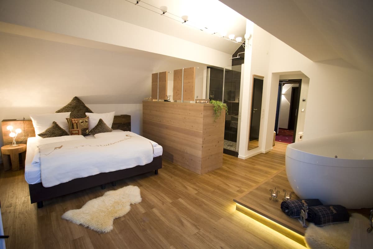 Un dormitorio moderno con una cama grande, una mesita de noche de madera, un tapete blanco, una bañera blanca amplia y un banco de madera con una planta.