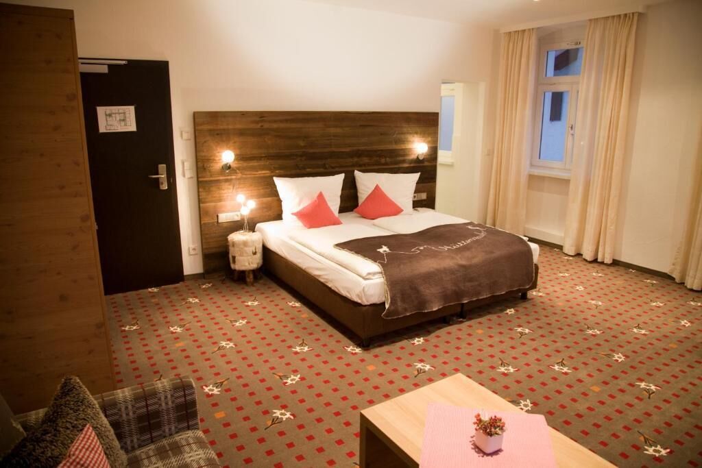 Ein Hotelzimmer mit hölzernem Kopfteil, einem Bett mit weißen Bettbezügen und roten Kissen, einem Nachttisch mit Lampe, einem kleinen Nachttisch mit Lampe, einem Fenster mit Vorhängen und einem gemusterten Teppich.