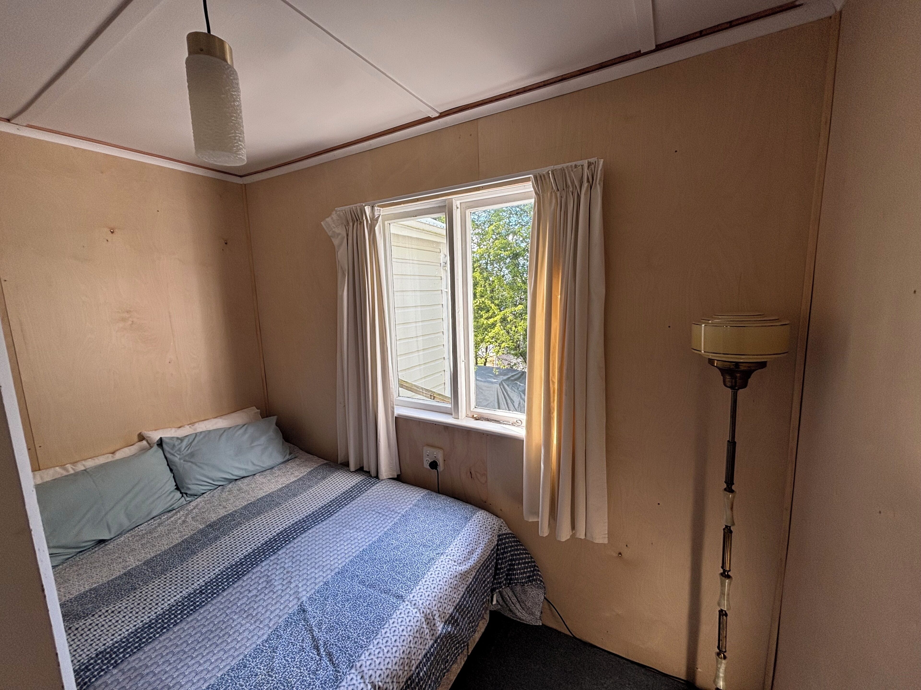 A bedroom with a bed, a window with curtains, a floor lamp, and a ceiling light.