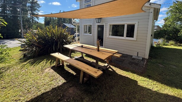 A wooden picnic table with benches, a shade sail, and a house with white siding and windows.