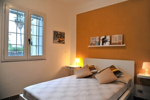 A neatly made bed with a patterned bedspread, a bedside lamp, and a small shelf with artwork.