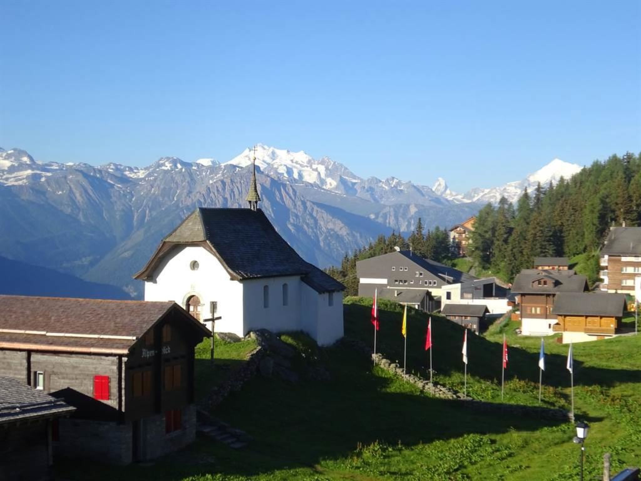 A small white church with a bell tower, surrounded by houses and flags, with a mountain range in the background.