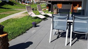 A terrace with lounge chairs and a table, overlooking a green landscape and buildings.