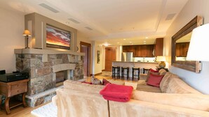 A living room with a stone fireplace, a sofa, and a kitchen in the background. - Big Horn Lodge 409 Delightful slopeside Residence (Truckee)