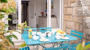 Une table à manger est dressée avec des bols et des tasses blancs, ainsi qu’une bouteille. Des chaises bleues l’entourent. On aperçoit en arrière-plan un mur de pierre et une porte donnant sur une cuisine.
