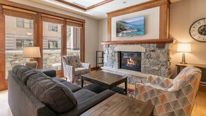 A cozy living room with a stone fireplace, comfortable seating, and a large window overlooking a stone wall. - Iron Horse Lodge North 206 - delightful slopeside (Truckee)