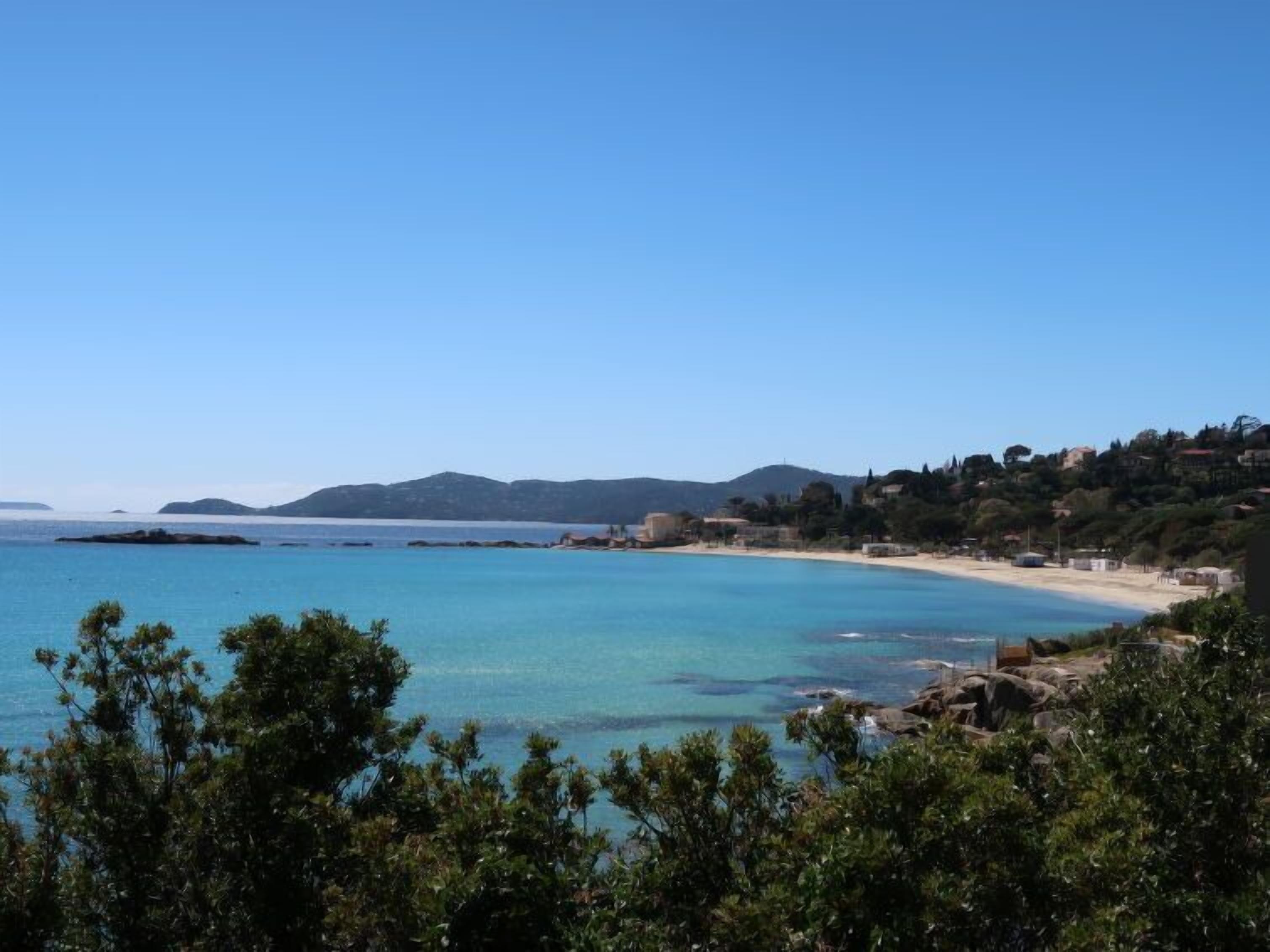A coastal view with clear blue waters, a sandy beach, and a hilly backdrop.