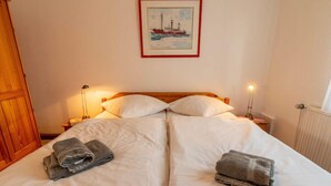 Ein Doppelbett mit weißen Bettwäsche, zwei Kissen und zwei Nachttischlampen. Darüber an der Wand hängt ein gerahmtes Bild.
