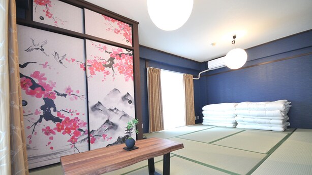 A traditional Japanese room with tatami flooring, a wooden table, a shoji screen with cherry blossom paintings, and a stack of white towels. - Maison Milano Nakatsu Apartment (Osaka)
