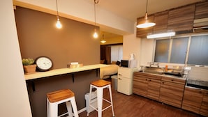 A compact kitchen with wooden cabinets, a microwave, a sink, and a clock on the wall. - Maison Milano Nakatsu Apartment (Osaka)