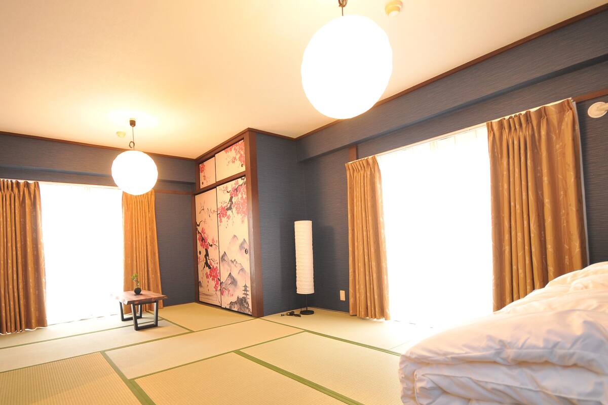 Habitación de hotel con suelo de tatami, una cama, un escritorio, una silla, una pintura tradicional y ventanas grandes con cortinas.