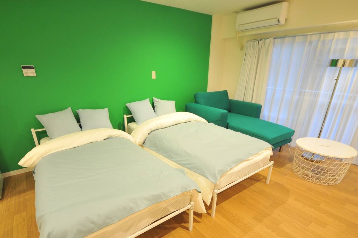 Una habitación con dos camas, una pared verde, un sofá, una mesita y una ventana con cortinas.