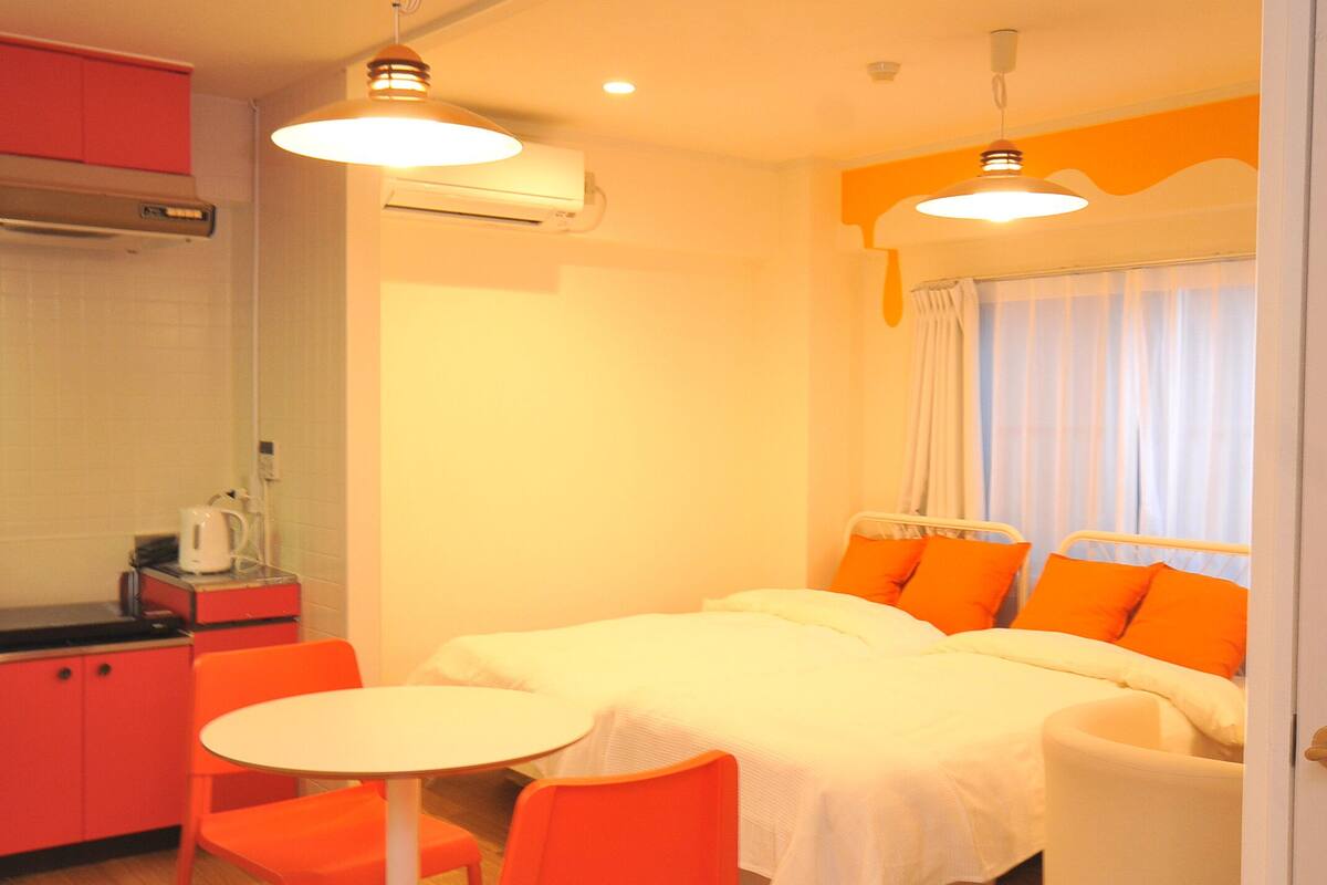 Habitación de hotel compacta con kitchenette, zona de comedor con mesa y sillas, y cama con almohadas naranjas.