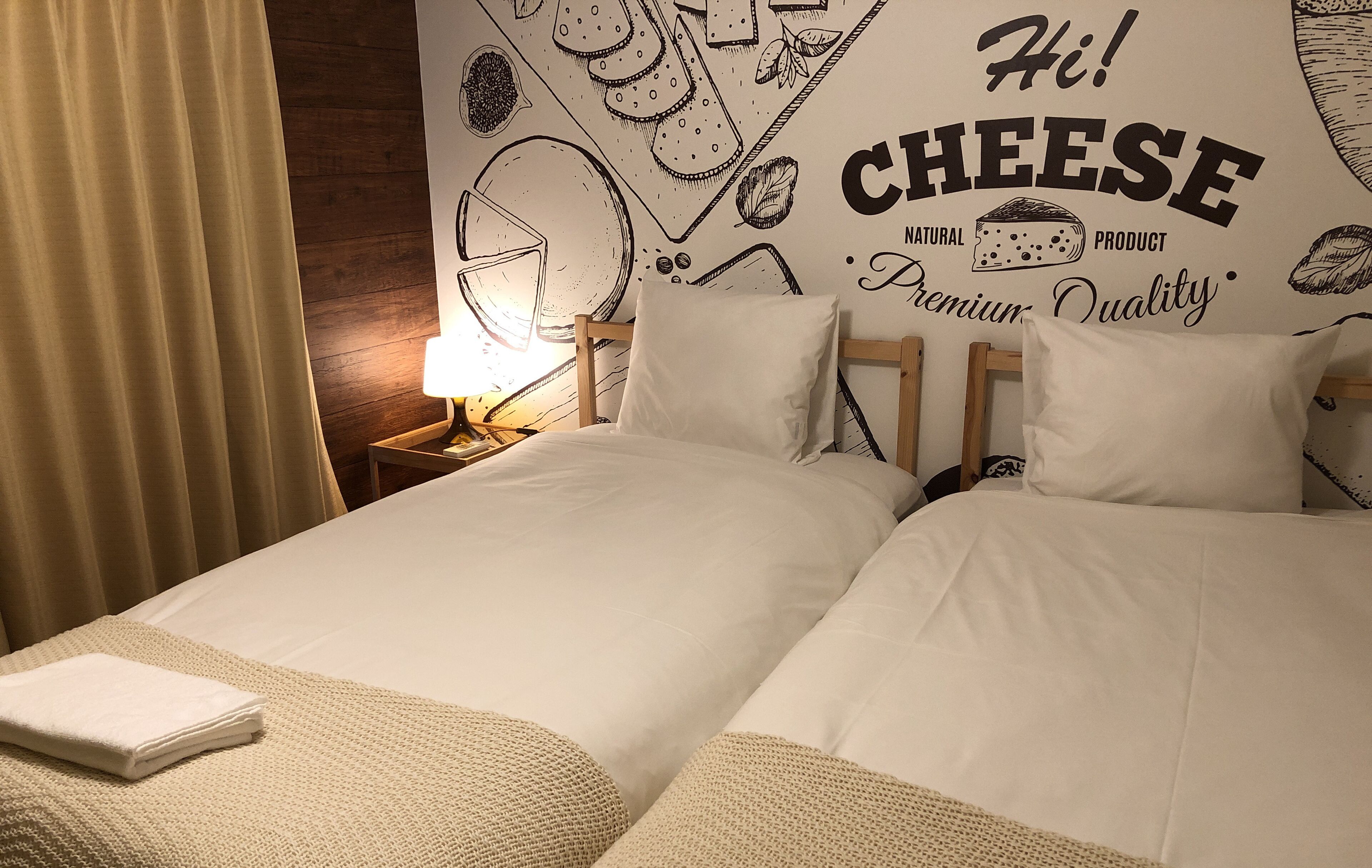 A hotel room with two beds, a wall mural with cheese illustrations, and a nightstand with a lamp.
