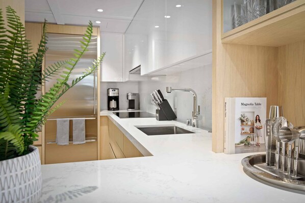 Una cocina moderna con encimera blanca, un lavabo y una cafetera. Hay una planta a la izquierda y una revista a la derecha.