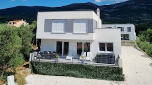 Una casa moderna de dos pisos con fachada blanca, un patio cubierto con mobiliario exterior y una vista de montañas al fondo.
