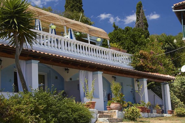 A two-story house with a balcony, surrounded by lush greenery and trees.
