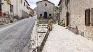 A narrow street with a stone building featuring a bell tower, a stone wall with a flower bed, and a paved sidewalk.