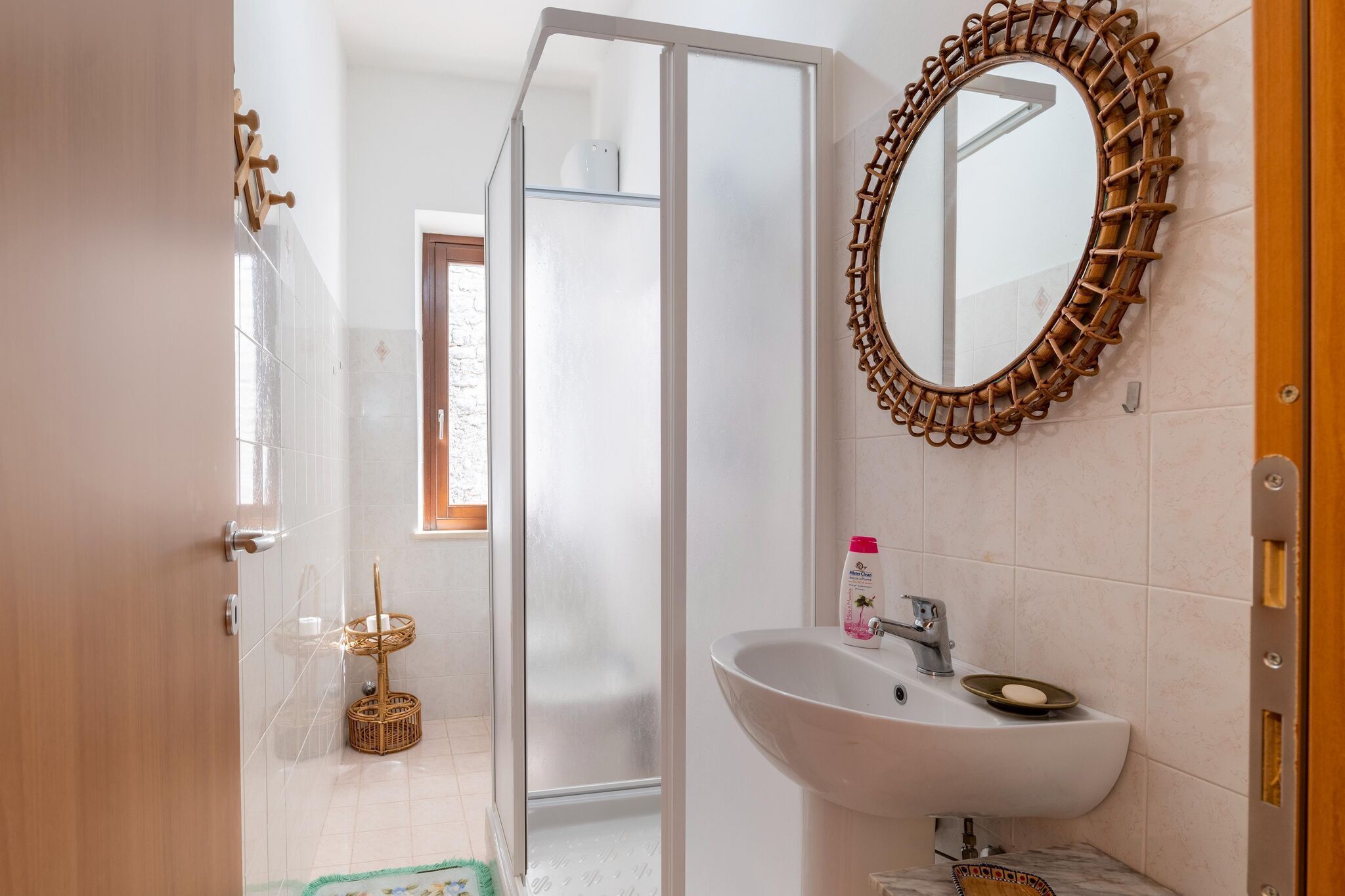 A bathroom with a glass shower door, a white sink with a soap dish, a wicker basket, and a mirror with a woven frame.