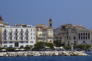 A coastal town with a prominent white building with balconies, a red-roofed tower, and a rocky shoreline with parked cars and umbrellas. - Amazing apartment in Mpenitses with terrace (Mpenitses)