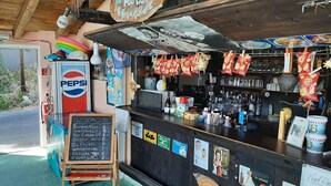 A poolside bar with a chalkboard menu listing snacks and drinks, a Pepsi refrigerator, and various snacks hanging above the bar.