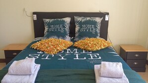 A neatly made bed with a green bedspread, two pillows with a tropical leaf pattern, and two folded towels.