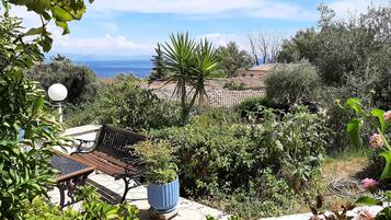 Een terras met een bankje en een plant in pot, omringd door weelderige begroeiing en uitzicht op zee.