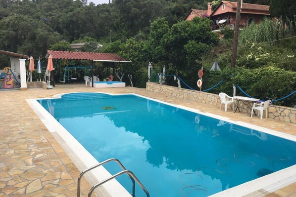Una piscina amb vora de pedra, una zona de jocs infantils i una zona de seients coberta amb ombrelles.
