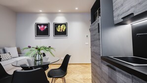 Una cocina moderna con un lavamanos integrado, un comedor con mesa de vidrio y sillas negras, y dos l áminas enmarcadas con motivos botánicos en la pared.