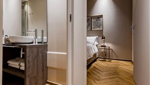Un baño moderno con un lavamanos grande, un espejo y una ducha. Junto a él, un dormitorio con cama, mesita de noche y lámpara de pie.