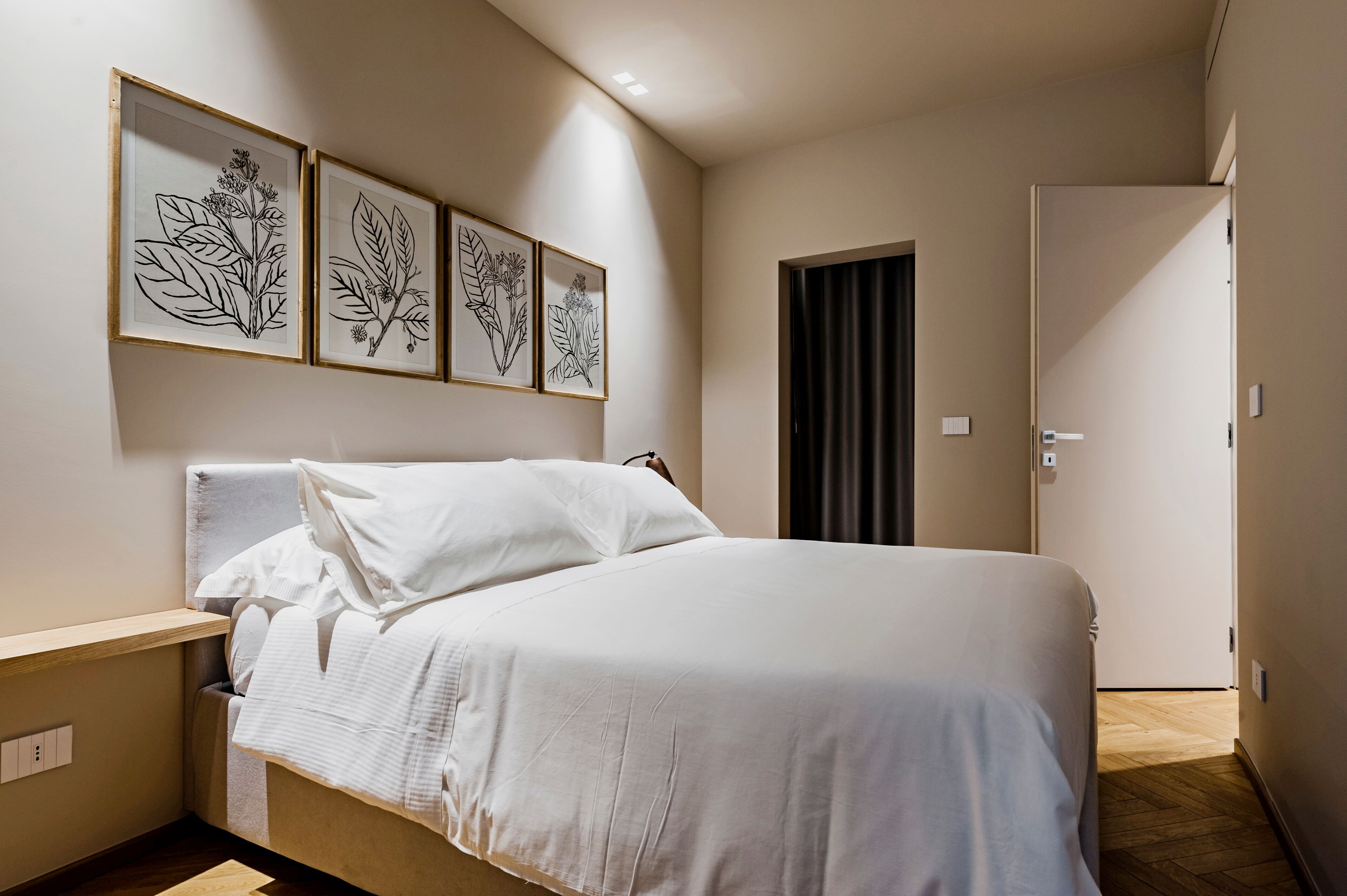 Una cama bien hecha con sábanas blancas en una habitación moderna con suelo de madera y cortina oscura en la puerta.