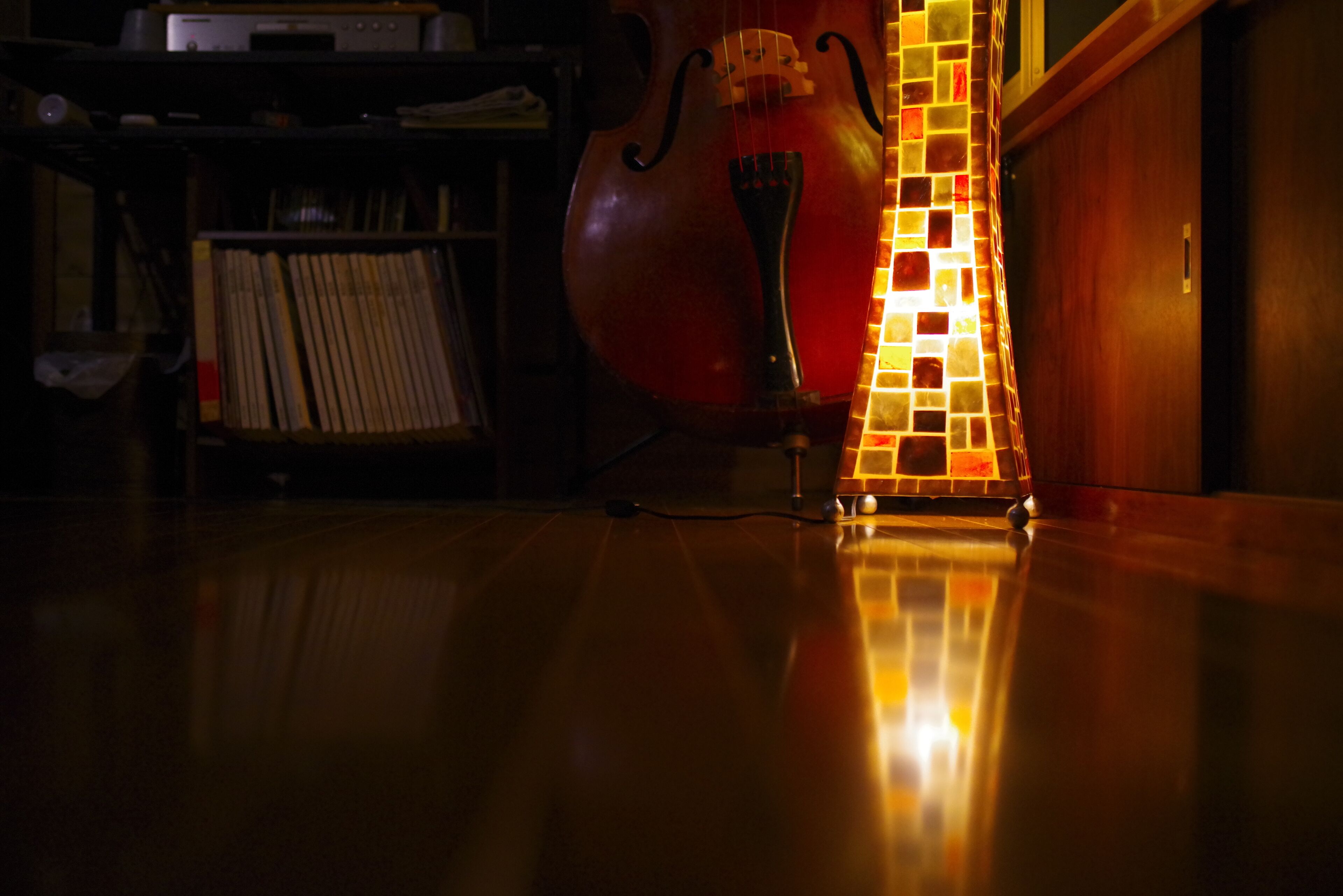 Een cello op een standaard, naast een mozaïeklamp in een zwak verlichte kamer met een houten vloer.