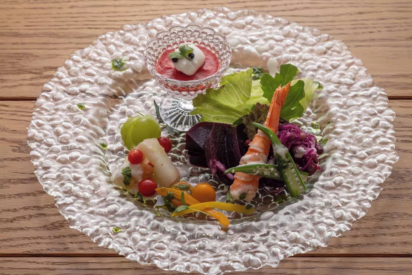 Prato de vidro arredondado e texturizado com diversos alimentos, incluindo frutos do mar, vegetais e um pequeno recipiente de vidro com uma porção de creme e uma folha verde para decorar.