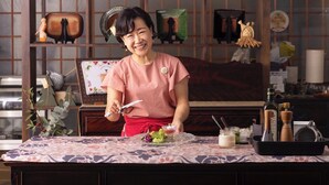 A person in a pink shirt holding a knife, standing at a table with a patterned tablecloth, surrounded by kitchen utensils and decorative items. - Azumino Salon (Azumino)