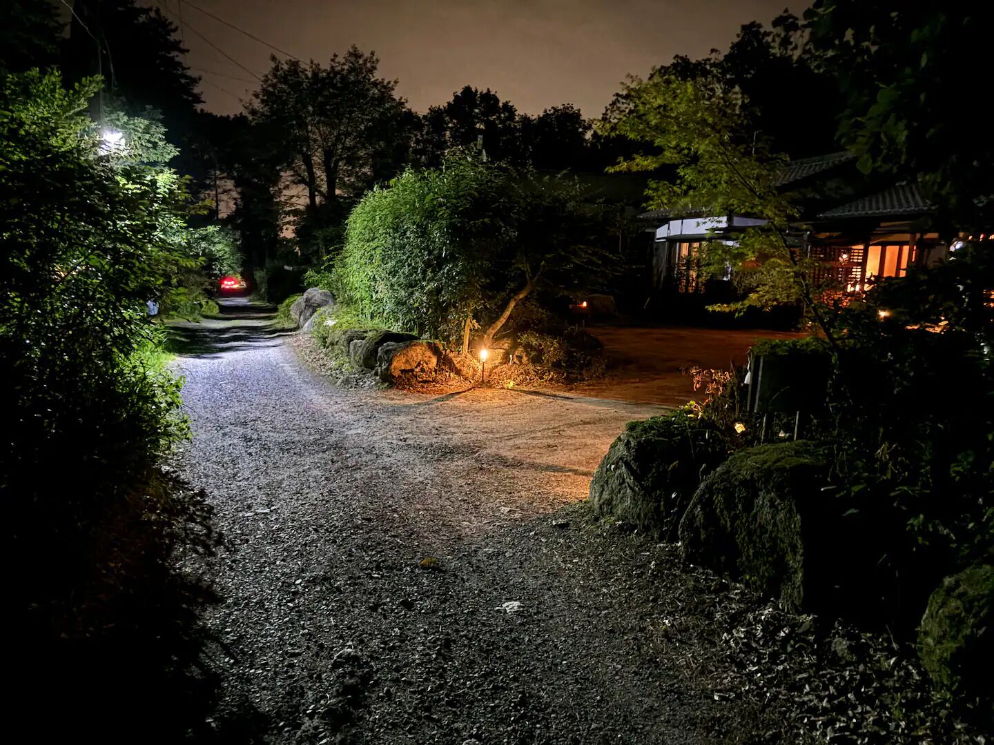 A gravel path leading to a traditional building with a tiled roof, surrounded by lush greenery and illuminated by street lamps.