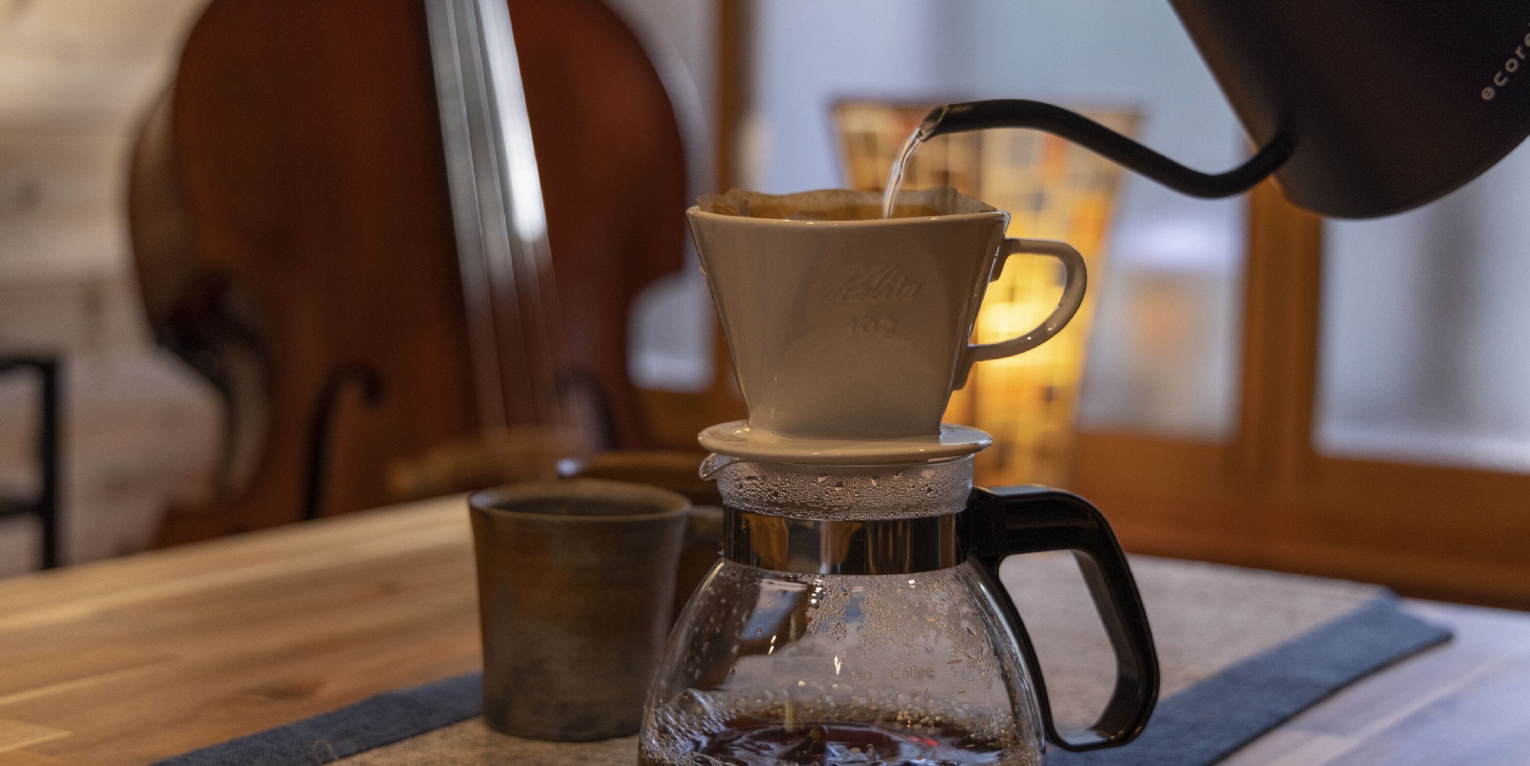 Uma cafeteira com jarra de vidro transparente e uma chávena de café branca sobre uma mesa de madeira.