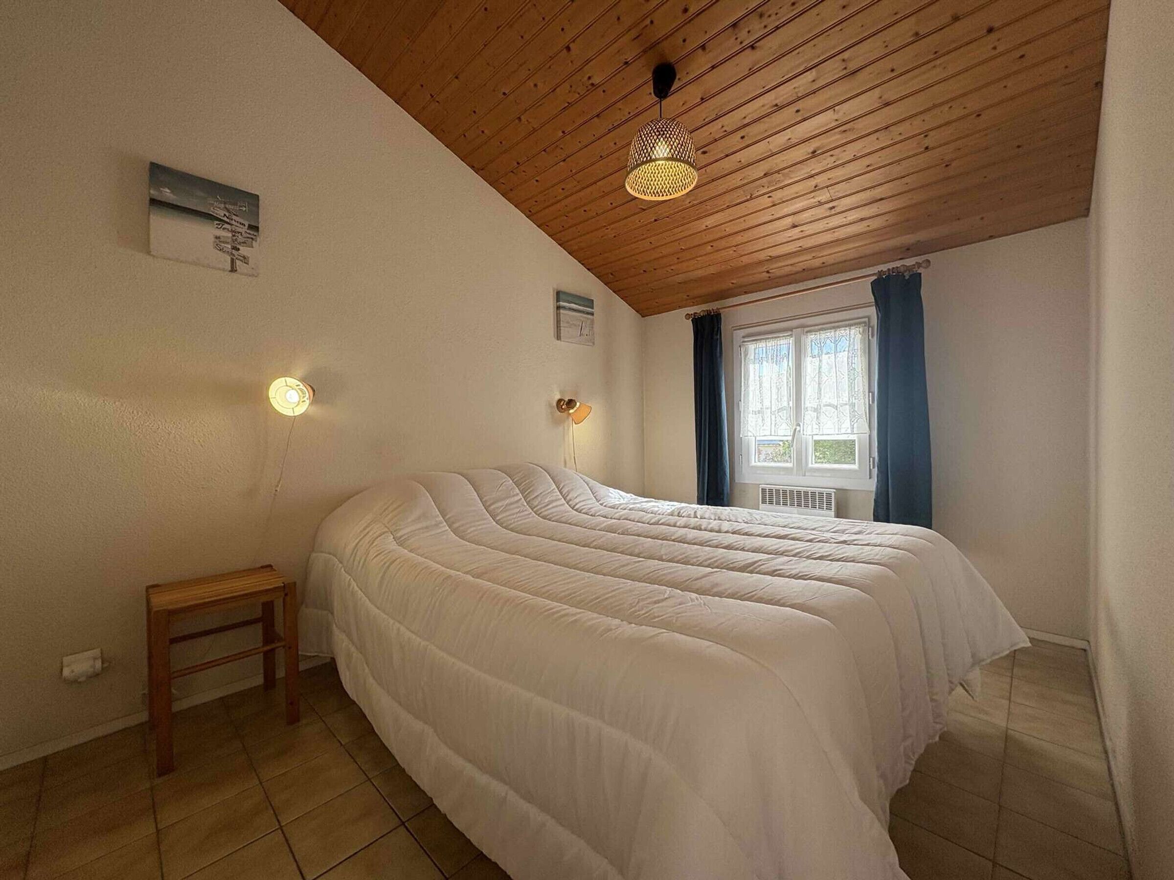 A bedroom with a wooden ceiling, a bed with a white duvet, a small wooden nightstand, a wall-mounted light, and a window with curtains.