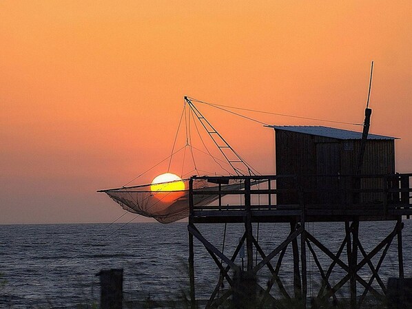 Un muelle con una red de pesca y una pequeña estructura de madera, posiblemente un mirador o depósito, con vista al mar durante la puesta de sol.