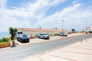 A coastal hotel with a red-tiled roof, a parking lot, and a view of the sea.