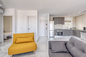 A modern apartment with a living room, a yellow sofa, a grey sofa, a kitchen with a built-in oven, and a white door.