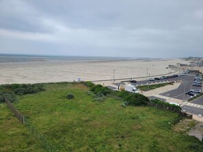 A coastal road with a beach in the background, a parking area, and a grassy area in the foreground.