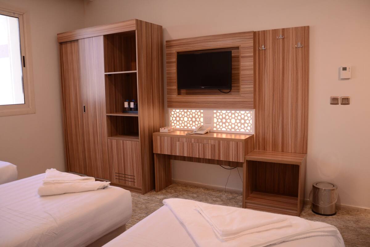 A hotel room with a wooden wardrobe, a desk with a flat-screen TV, a vanity with a mirror, and two beds.