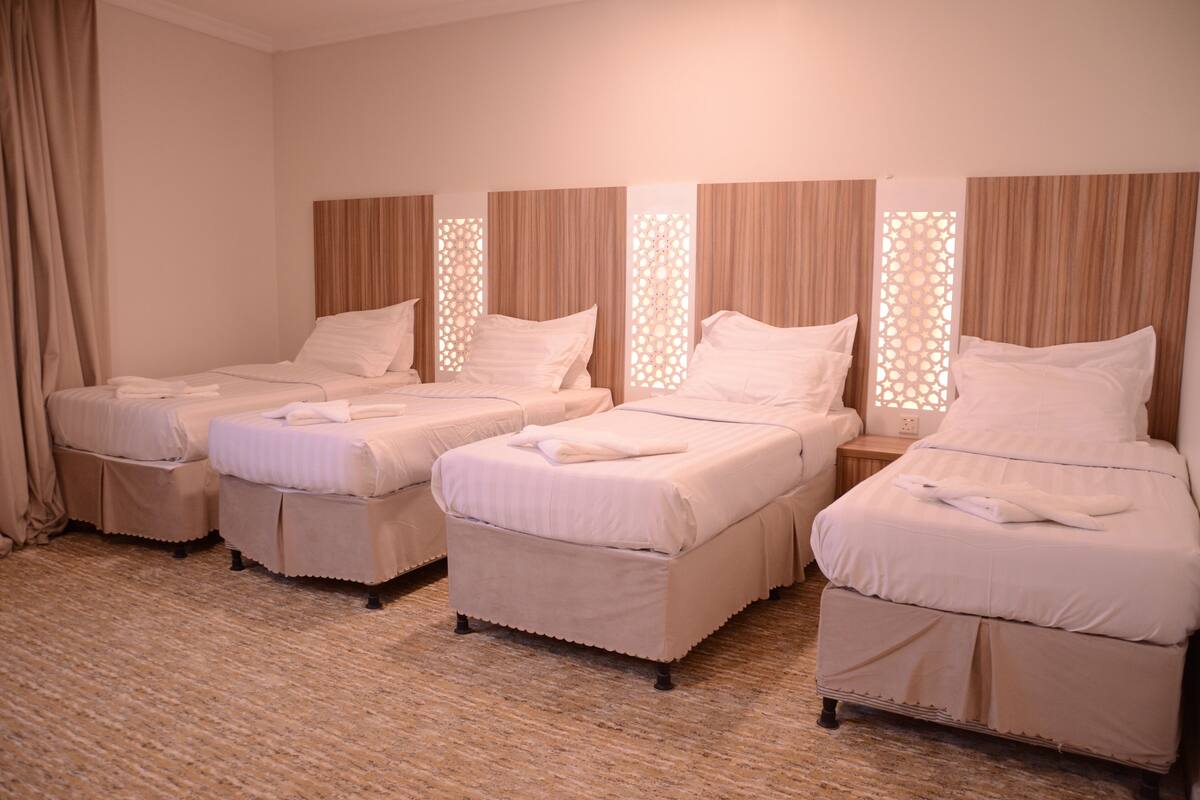 A hotel room with three single beds, each with a white bedspread and pillows, and a small bedside table with a lamp.