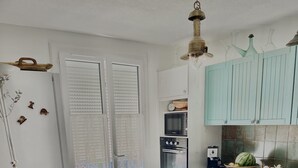 A kitchen with white cabinets, a microwave, oven, and a dishwasher. There is a dining table with a chair and a window with blinds. - Charming, bright house 10 minutes from the village center (Collioure)