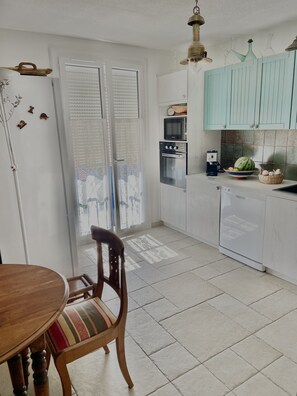 A kitchen with white cabinets, a microwave, oven, and a dishwasher. There is a dining table with a chair and a window with blinds.