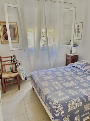 A bedroom with a bed, a chair, a nightstand, a lamp, and a window with sheer curtains.