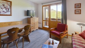 Una habitación con una mesa de comedor de madera, sillas, un sofá rojo, un televisor sobre un mueble de madera y una ventana con vistas a un paisaje verde.