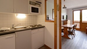 Cocina compacta con muebles blancos, microondas, lavaplatos y fregadero. En el área de comedor contigua, hay una mesa de madera con sillas.
