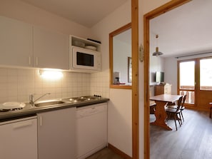 A compact kitchen with white cabinets, a microwave, a sink, and a dishwasher. There is a wooden dining table with chairs in the adjacent dining area.