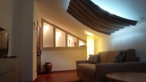 A living room with a sofa, a small table, and a television. There is a window with curtains, a vase on the floor, and a wall-mounted shelf with books. - Attic apartment iPatrizi (Moncalieri)
